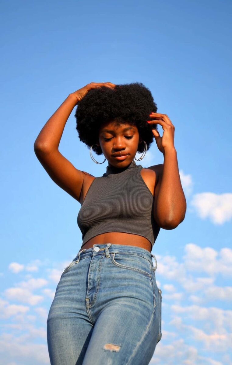 A Young Black Woman In A Crop Top And Jeans Posing In Front Of A Blue Sky.