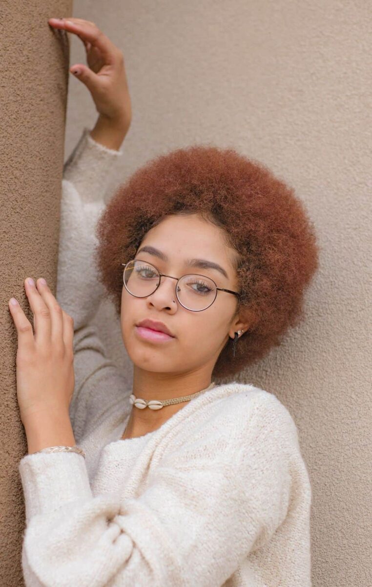 A Young Woman Wearing Glasses And A Sweater Leaning Against A Wall.