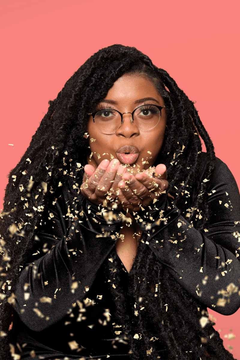 A Woman With Dreadlocks Blowing Out Gold Confetti.