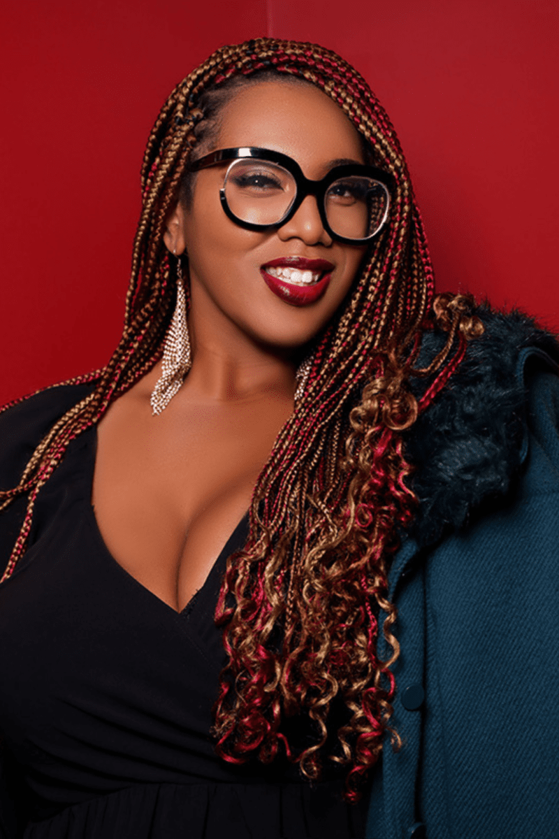A Black Woman With Glasses And Braids Posing For A Photo.