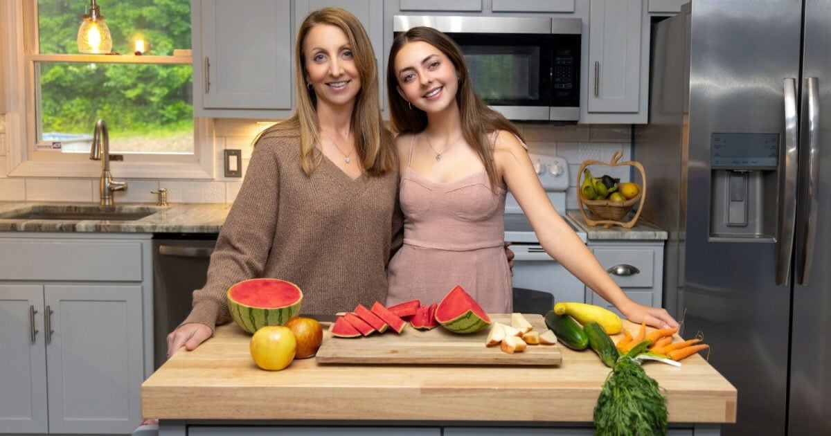 Two Women Posing In Front Of A Cutting Board With Fruits And Vegetables For Pictures.