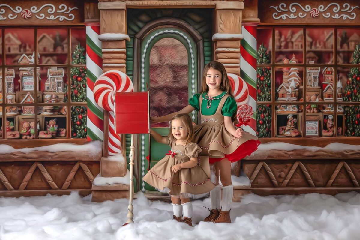 Two Little Girls Excitedly Pose In Front Of A Charming Gingerbread House During Christmas Mini Sessions.
