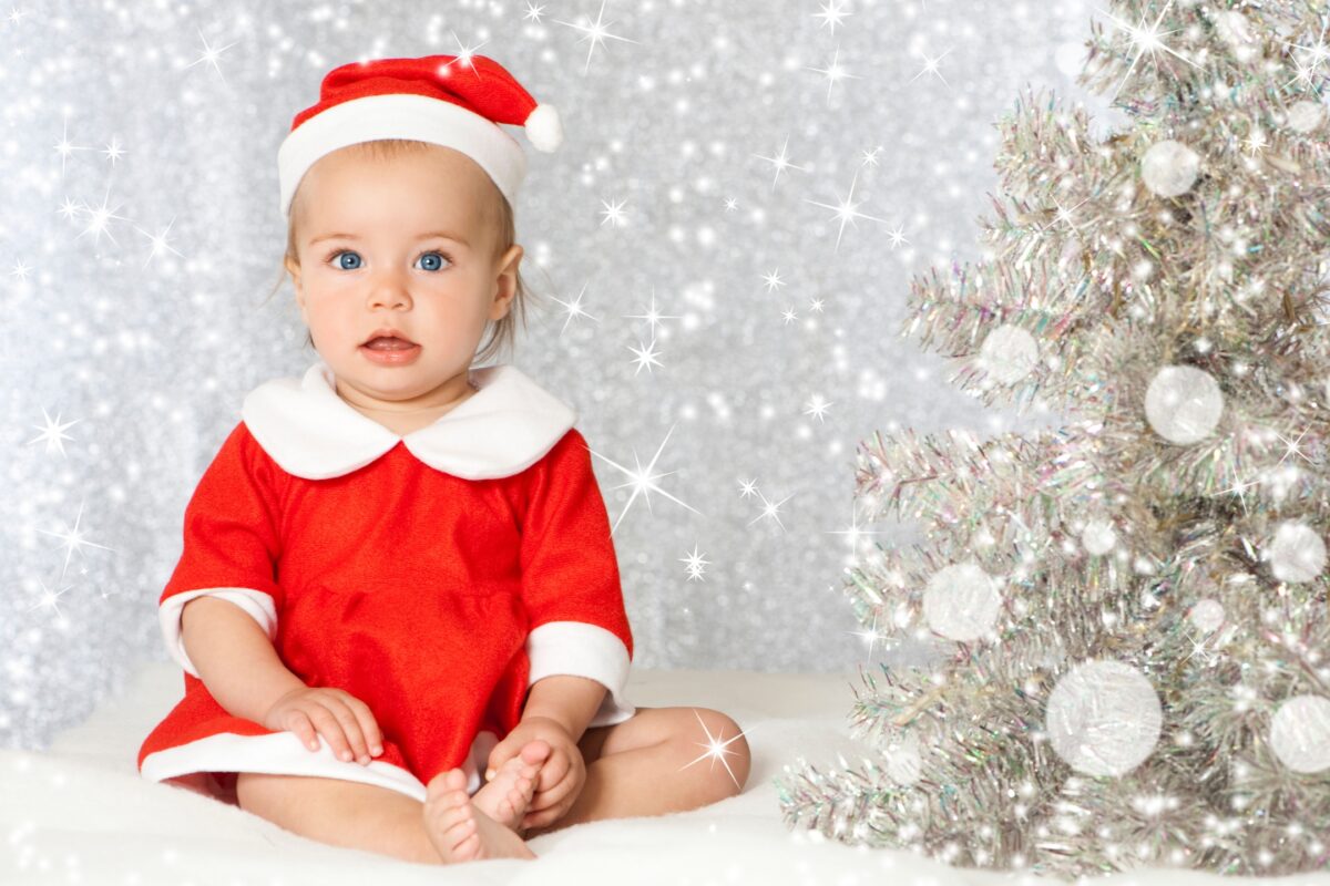 A Baby In A Santa Claus Costume Poses For A Santa Photoshoot In Front Of A Christmas Tree.