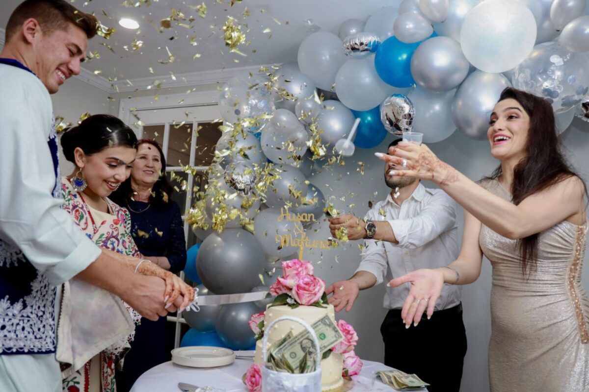 A Group Of People Cutting A Cake In Front Of Balloons.