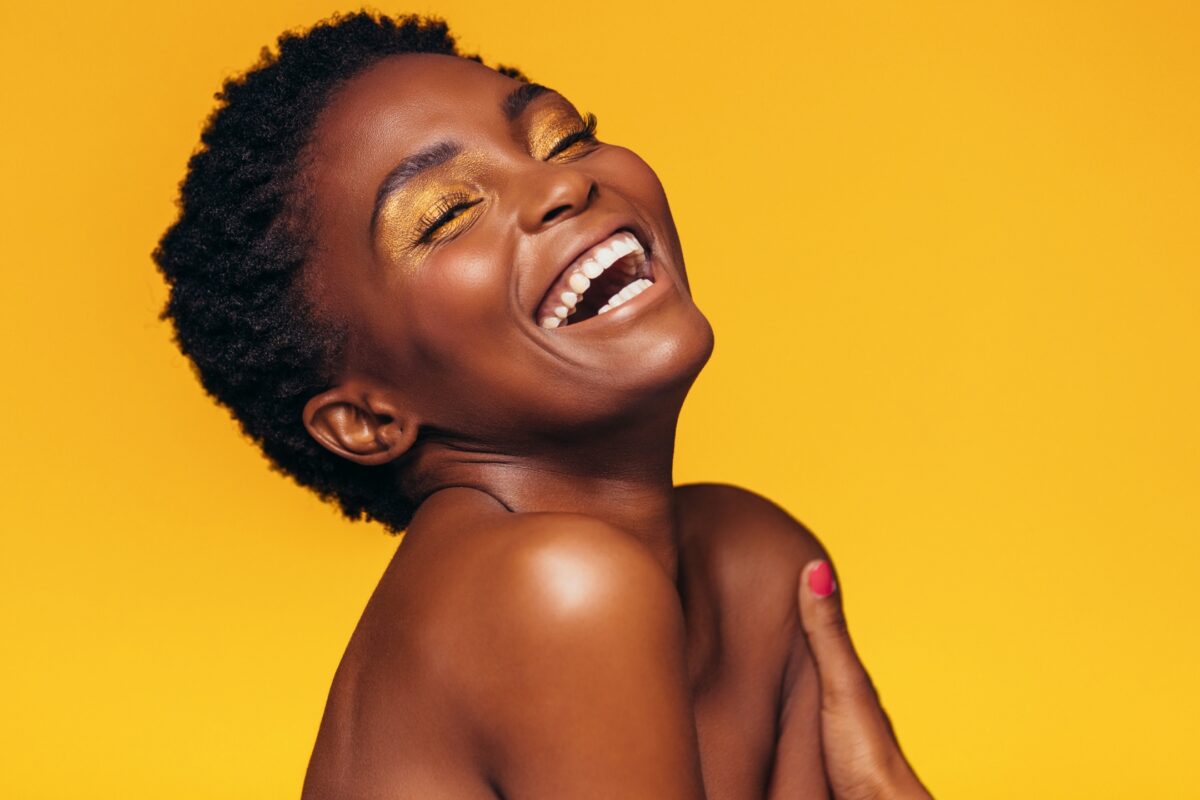 A Young Black Woman Laughing On A Vibrant Yellow Background, Guided By Posing Prompts During The Photo Session.