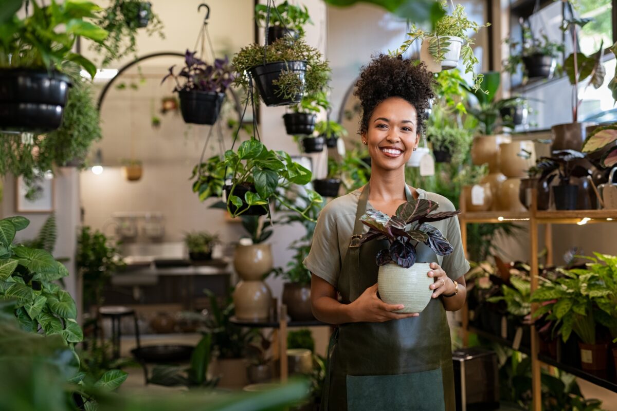 A Woman In An Apron Is Holding A Potted Plant In A Store, Showcasing Why Photography Is Important.