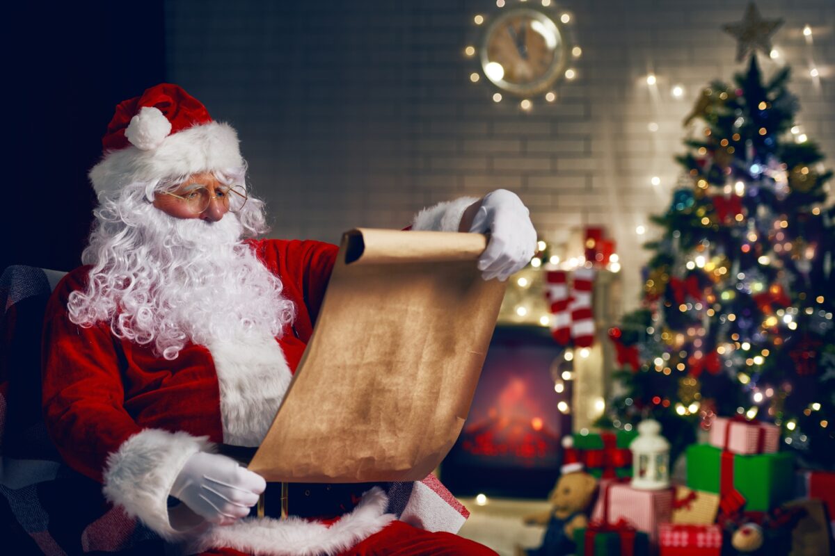 Santa Claus Posing For A Santa Photoshoot, Holding A Scroll In Front Of A Christmas Tree.
