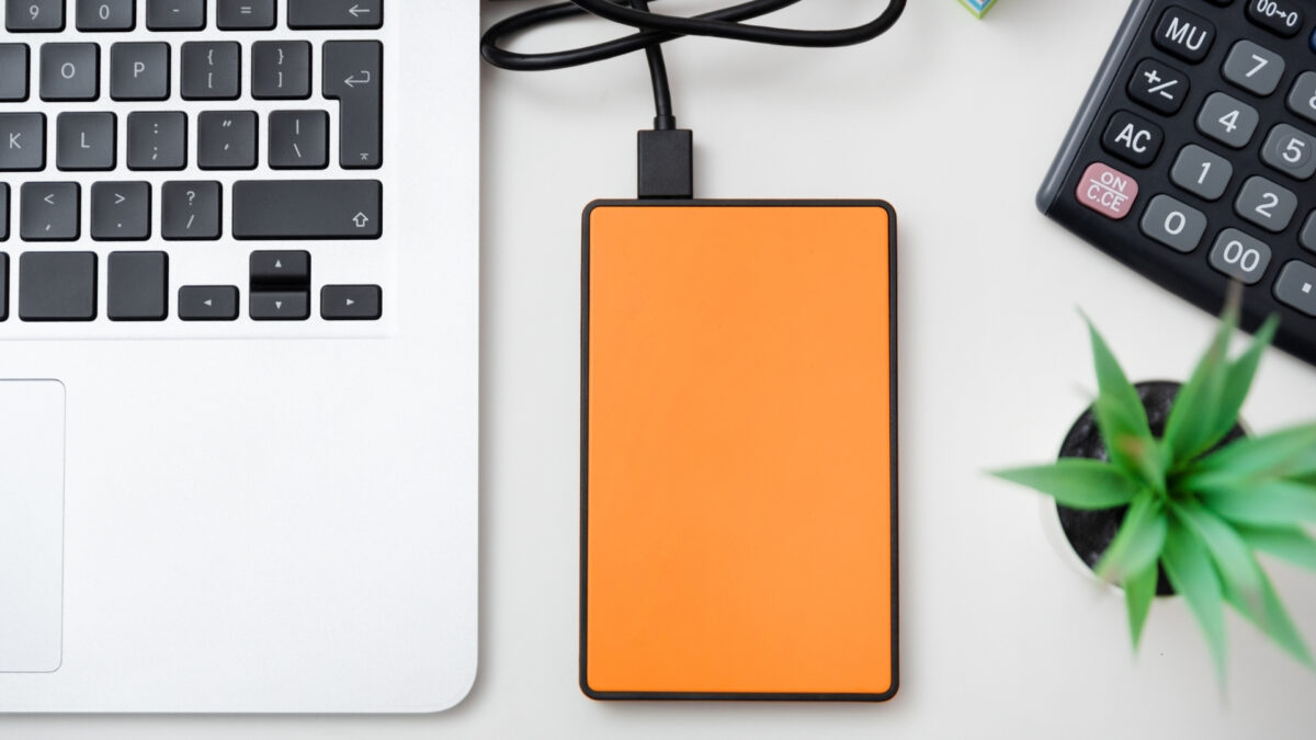 An Orange Power Bank Conveniently Placed Next To A Laptop And A Plant, Providing The Perfect Portable Solution To Store And Backup Photos On-The-Go.
