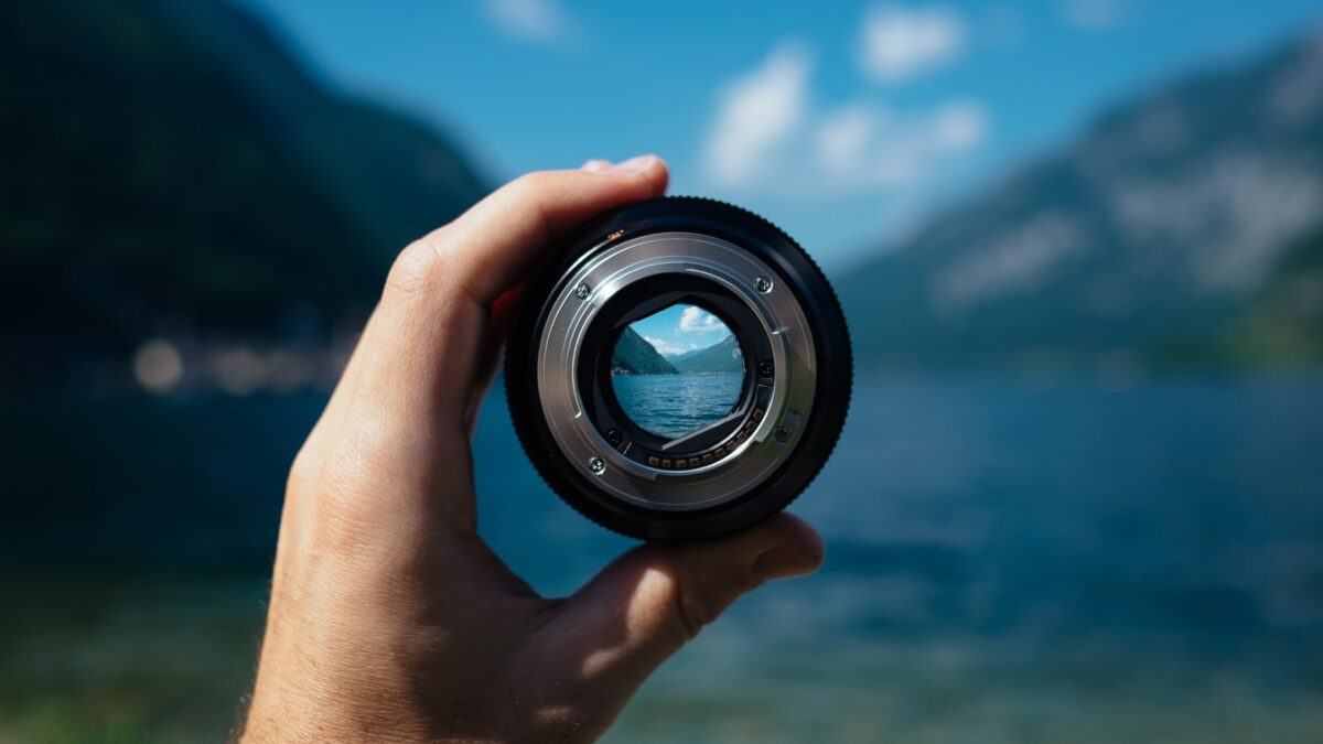 A Person Making Mistakes As A Photographer While Holding Up A Camera Lens With A Lake In The Background.