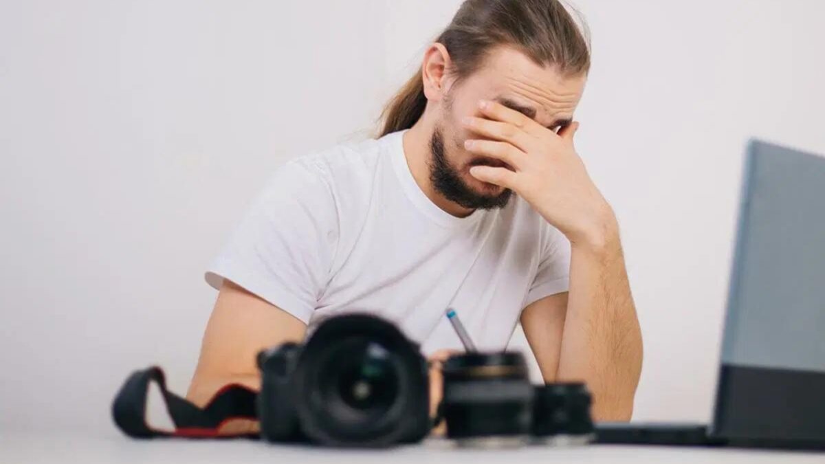 A Man Covering His Face In Frustration While Sitting At A Desk With A Laptop, Reflecting On His Mistakes As A Photographer.