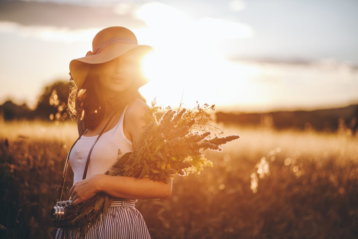 A Woman In A Hat Holding Flowers In A Field During Sunset, Captured With The Warm Glow Of Harsh Sunlight.