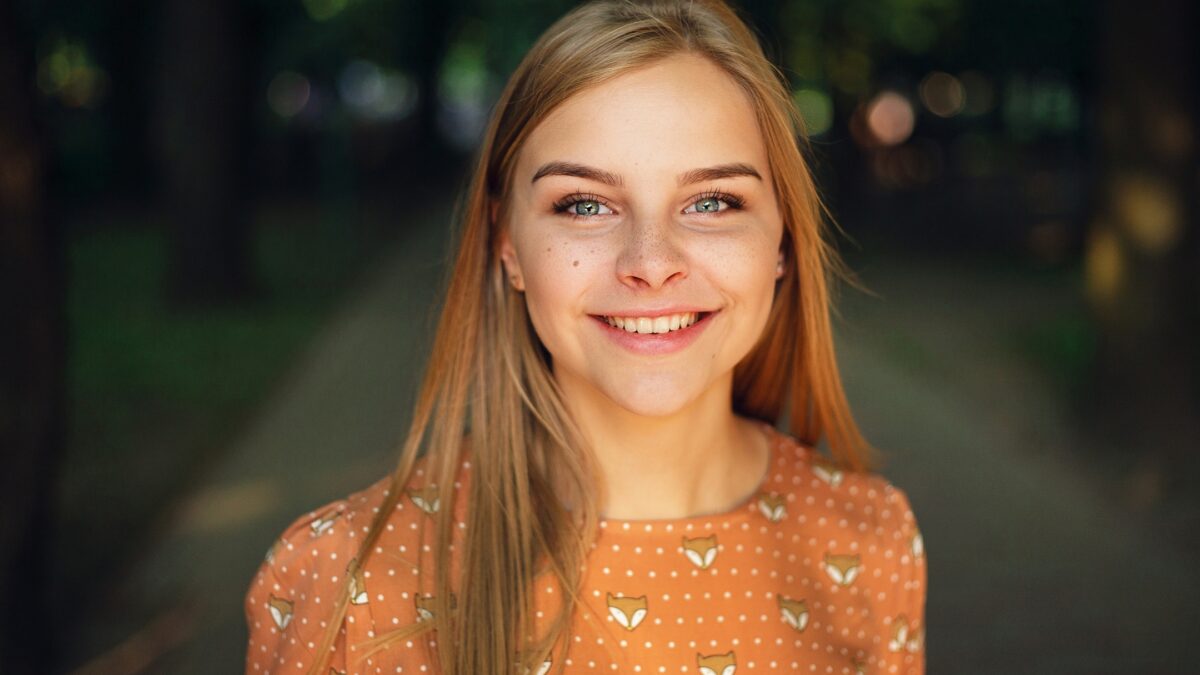 A Young Woman Is Smiling In A Park While A Photography Lighting Setup Captures Her Joy.