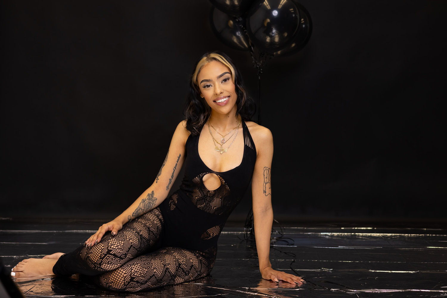 Connecticut A Woman In A Black, Patterned Outfit Sits On A Shiny Black Floor With Black Balloons In The Background, Smiling At The Camera, Showcasing Our Event Styling Services.