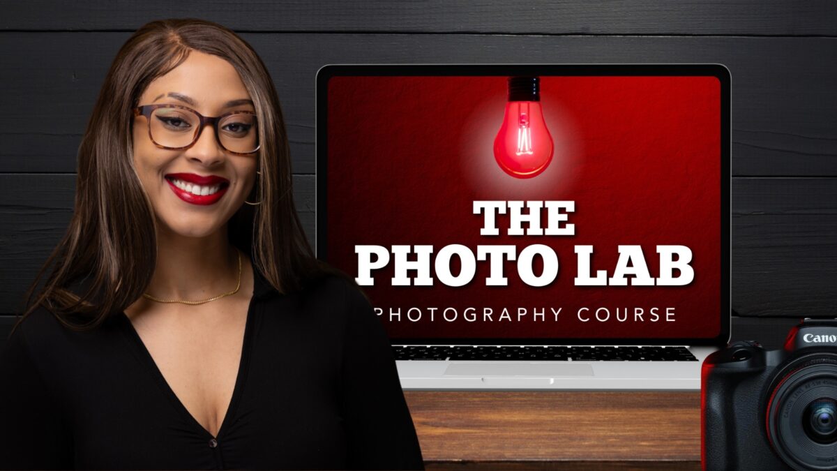 Smiling Woman With Glasses Facing The Camera, Standing In Front Of A Laptop Displaying &Quot;The Photo Lab Video Course&Quot; And A Red Light Bulb Image; Camera Partially Visible In The Foreground.